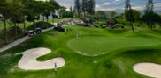 Green between curved bunkers at Barcelona Valley Lake Course, with a rocky outcrop, tall trees and a cart path alongside.