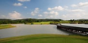 Curving bridge across water at Barcelona Valley Mountain Course, with grassy banks, a fairway and wooded hills beyond.