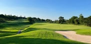 Green beside a curved sand bunker, with an undulating fairway and trees beyond.