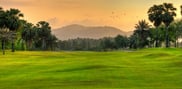 The palm-lined 18th fairway beneath a golden sky, with bunkers and distant hills.
